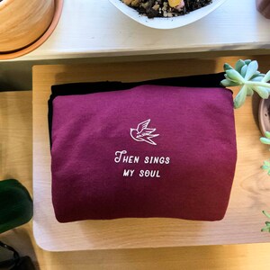 Puede incluir: Sudadera color burdeos con un p&aacute;jaro bordado en blanco y el texto "Then Sings My Soul".