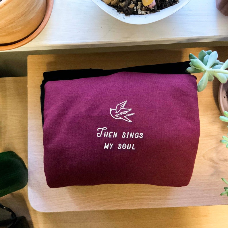 Puede incluir: Sudadera color burdeos con un p&aacute;jaro bordado en blanco y el texto "Then Sings My Soul".