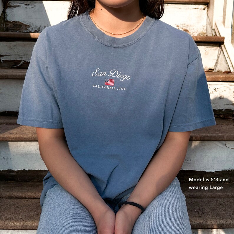May include: A light blue t-shirt with the text "San Diego, California, USA" and a small embroidered American flag. The model is 5'3" and wearing a size Large.