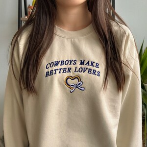 May include: A cream colored sweatshirt with the text "COWBOYS MAKE BETTER LOVERS" embroidered in blue and yellow thread. A heart shape is embroidered below the text.