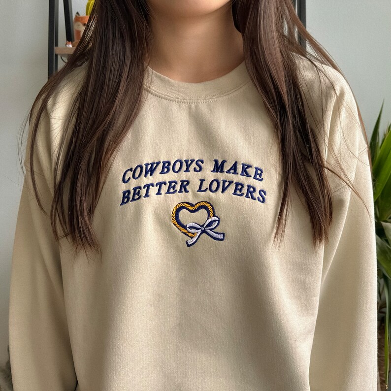 May include: A cream colored sweatshirt with the text "COWBOYS MAKE BETTER LOVERS" embroidered in blue and yellow thread. A heart shape is embroidered below the text.