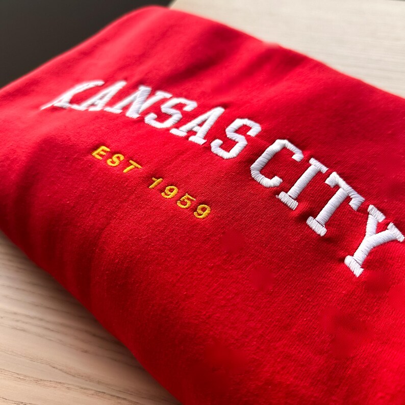 Puede incluir: Una sudadera roja doblada con el texto bordado "KANSAS CITY" en blanco, arqueado en la parte superior. Debajo, en hilo dorado, est&aacute; "EST 1959". La tela es de un rojo s&oacute;lido y vibrante.