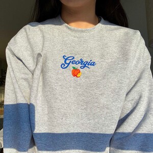 K&ouml;nnte beinhalten: Grauer Pullover mit einer gestickten blauen und orangenen Pfirsich und dem Wort "Georgia" auf der Vorderseite. Der Pullover hat einen blauen Streifen am unteren Rand.