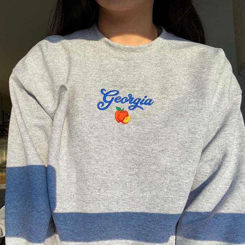 K&ouml;nnte beinhalten: Grauer Pullover mit einer gestickten blauen und orangenen Pfirsich und dem Wort "Georgia" auf der Vorderseite. Der Pullover hat einen blauen Streifen am unteren Rand.
