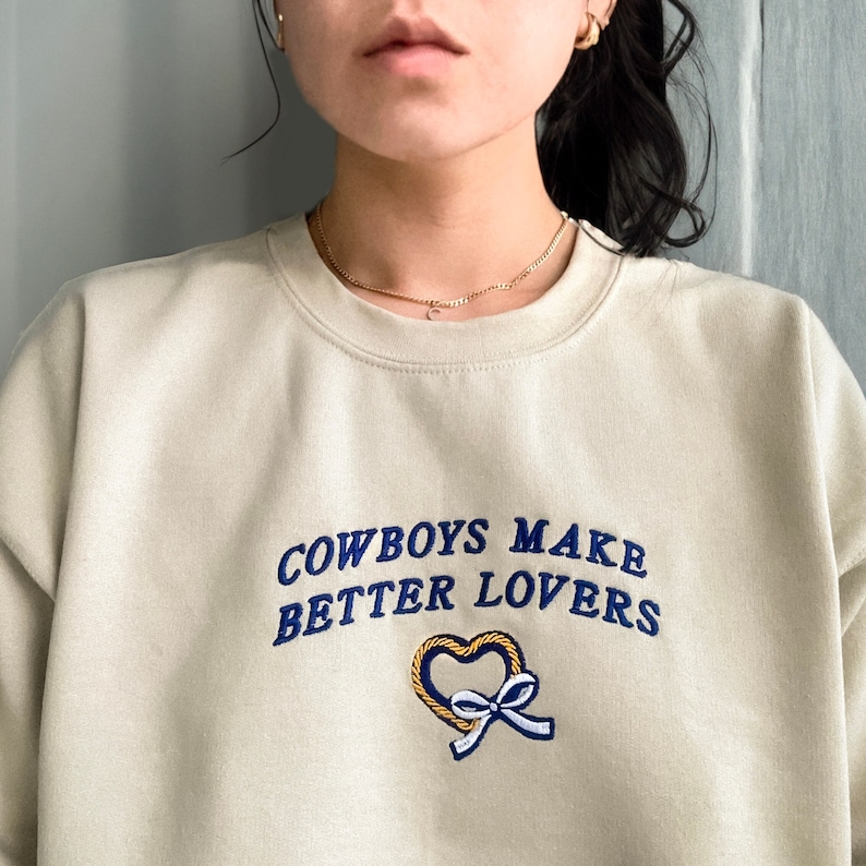 May include: A light beige sweatshirt with the text "COWBOYS MAKE BETTER LOVERS" in blue embroidery. A heart with a bow is embroidered below the text.