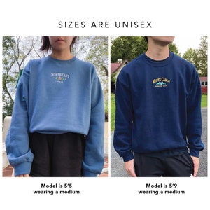 Op de afbeelding: Twee modellen dragen blauwe crewneck sweatshirts met geborduurde logo's. De trui aan de linkerkant is lichtblauw met de tekst "Northeast League" en de trui aan de rechterkant is donkerblauw met de tekst "Monte Carlo Country Club".