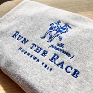 May include: A light gray sweatshirt with blue embroidered text and a running figure design. The text reads "RUN THE RACE" with "Hebrews 12:1" and "with perseverance!" below the main text. The design features two runners.