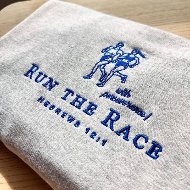 May include: A light gray sweatshirt with blue embroidered text and a running figure design. The text reads "RUN THE RACE" with "Hebrews 12:1" and "with perseverance!" below the main text. The design features two runners.