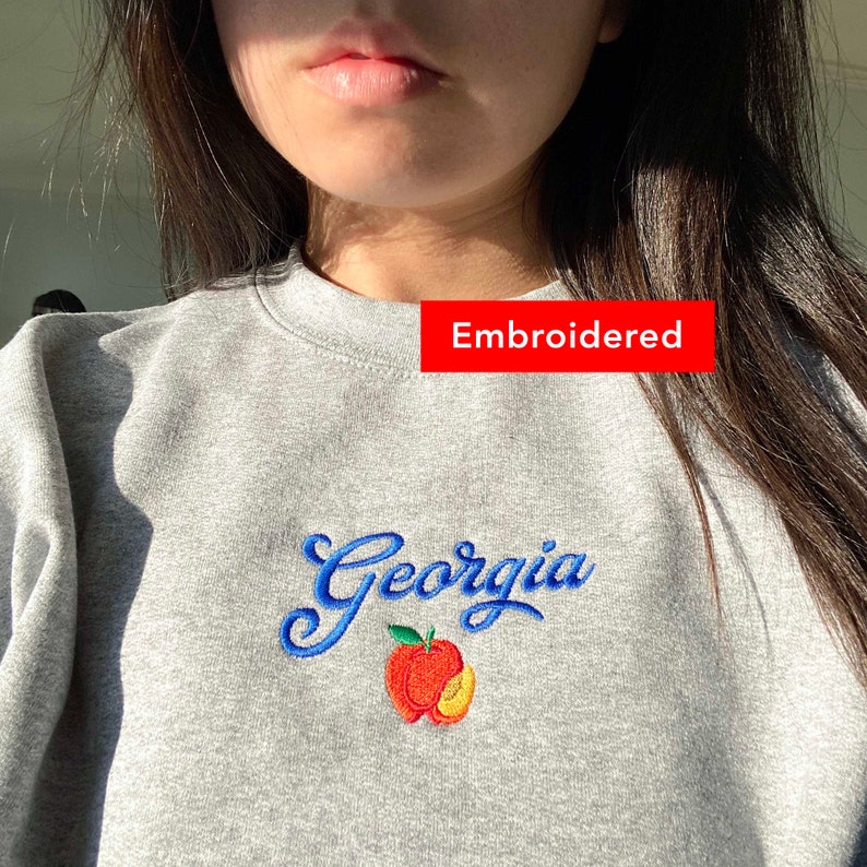 K&ouml;nnte beinhalten: Grauer Sweatshirt mit blau gesticktem Text "Georgia" und einer Pfirsichgrafik.