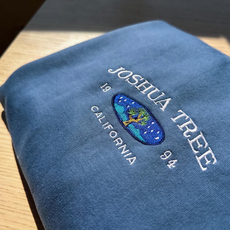May include: A blue sweatshirt with an embroidered logo that says "Joshua Tree 1994 California". The logo features a stylized Joshua tree in a blue oval with white stars.