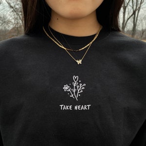 May include: Black long-sleeve shirt with a white embroidered design of a flower with a heart and the text "TAKE HEART". The model is wearing a gold chain necklace with a butterfly pendant.