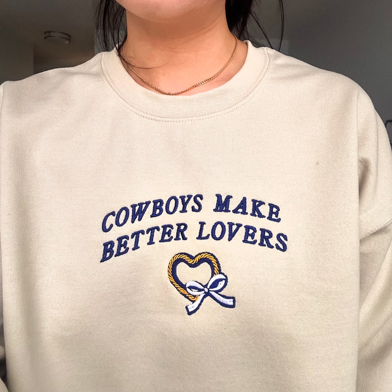 May include: A cream-colored sweatshirt with the text "COWBOYS MAKE BETTER LOVERS" in navy blue embroidery. A gold heart with a navy blue bow is embroidered below the text.