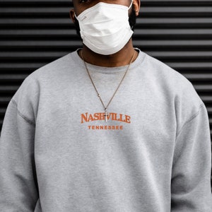 May include: A gray sweatshirt with the text "NASHVILLE TENNESSEE" in orange lettering. The sweatshirt has a silver chain necklace with a small cross pendant.