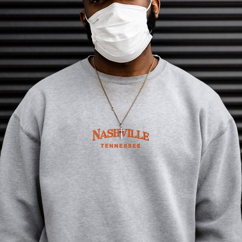 May include: A gray sweatshirt with the text "NASHVILLE TENNESSEE" in orange lettering. The sweatshirt has a silver chain necklace with a small cross pendant.