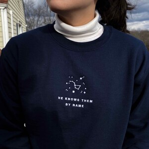 Puede incluir: Una sudadera azul marino con una constelaci&oacute;n bordada en blanco y el texto "HE KNOWS THEM BY NAME".