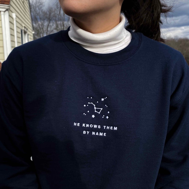 Puede incluir: Una sudadera azul marino con una constelaci&oacute;n bordada en blanco y el texto "HE KNOWS THEM BY NAME".