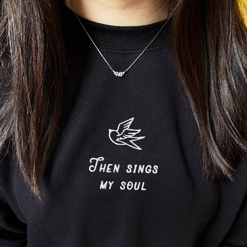 Puede incluir: Sudadera negra con un p&aacute;jaro bordado en blanco y el texto "Then Sings My Soul". La sudadera tiene un delicado collar de plata con un peque&ntilde;o colgante.
