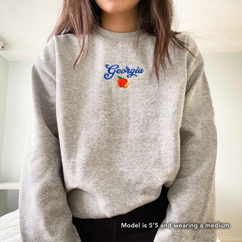 K&ouml;nnte beinhalten: Grauer Sweatshirt mit einem blauen gestickten Design, das "Georgia" mit einer Pfirsichgrafik sagt.