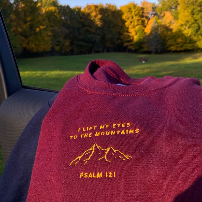May include: A maroon sweatshirt with the text "I lift my eyes to the mountains" and "PSALM 121" embroidered in yellow. The mountains are also embroidered in yellow.