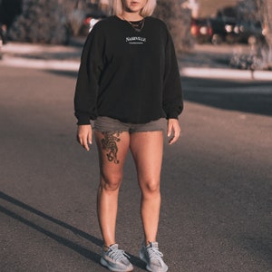 May include: A woman wearing a black sweatshirt with the text "Nashville Tennessee" on the front, denim shorts, and white sneakers. She has a tiger tattoo on her left thigh.