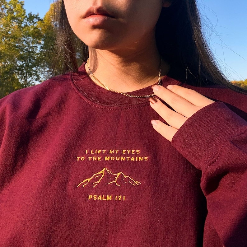 May include: Burgundy sweatshirt with a gold embroidered mountain design and the text "I lift my eyes to the mountains Psalm 121".