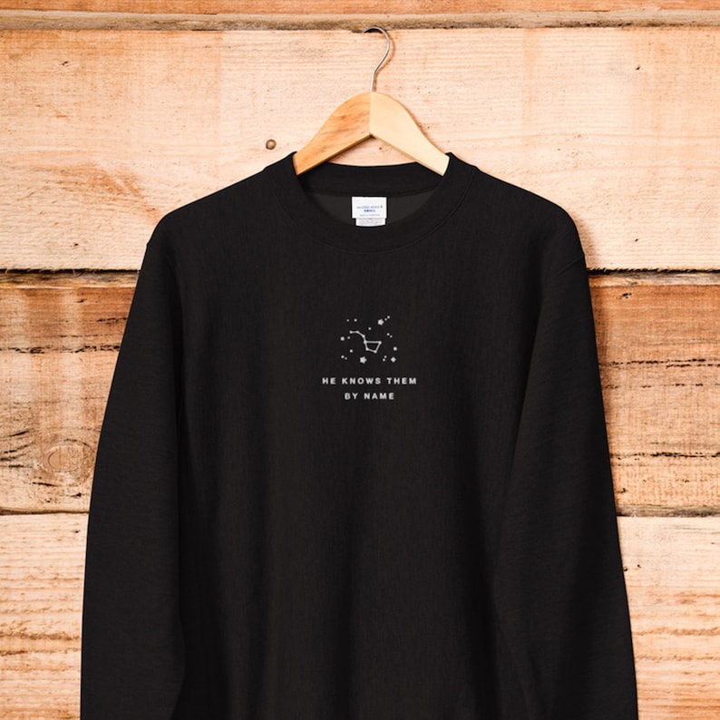 Puede incluir: Sudadera negra de manga larga con un gr&aacute;fico de constelaci&oacute;n blanca y el texto "HE KNOWS THEM BY NAME".