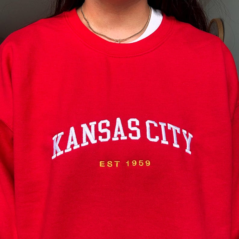 Puede incluir: Una sudadera roja con el texto blanco "KANSAS CITY" en arco. Debajo, el texto dorado dice "EST 1959". La sudadera es de color rojo liso.