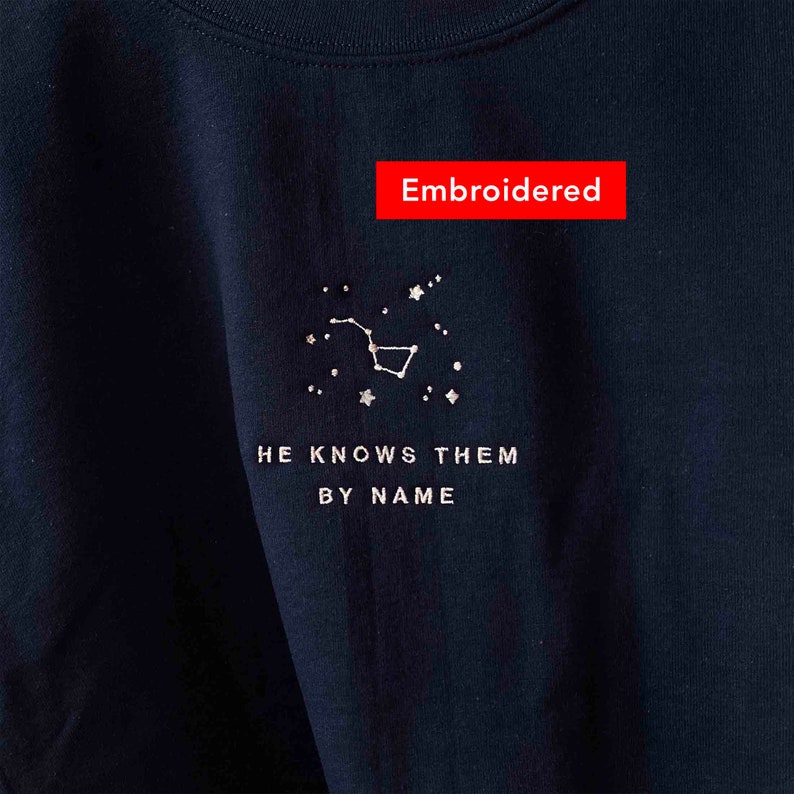 Puede incluir: Una camiseta negra con un dise&ntilde;o de constelaci&oacute;n bordado y el texto "HE KNOWS THEM BY NAME". El bordado es plateado.