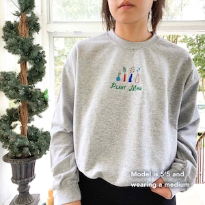 May include: Gray sweatshirt with a green, red, blue, and white embroidered design of plants in pots. The text "PLANT MOM" is embroidered below the design.