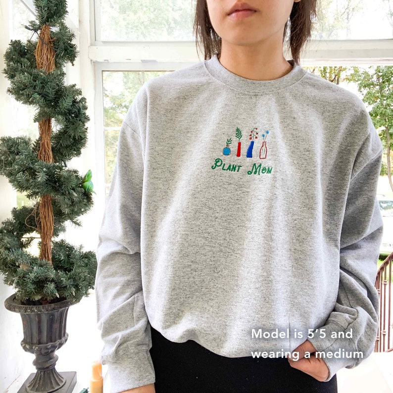 May include: Gray sweatshirt with a green, red, blue, and white embroidered design of plants in pots. The text "PLANT MOM" is embroidered below the design.