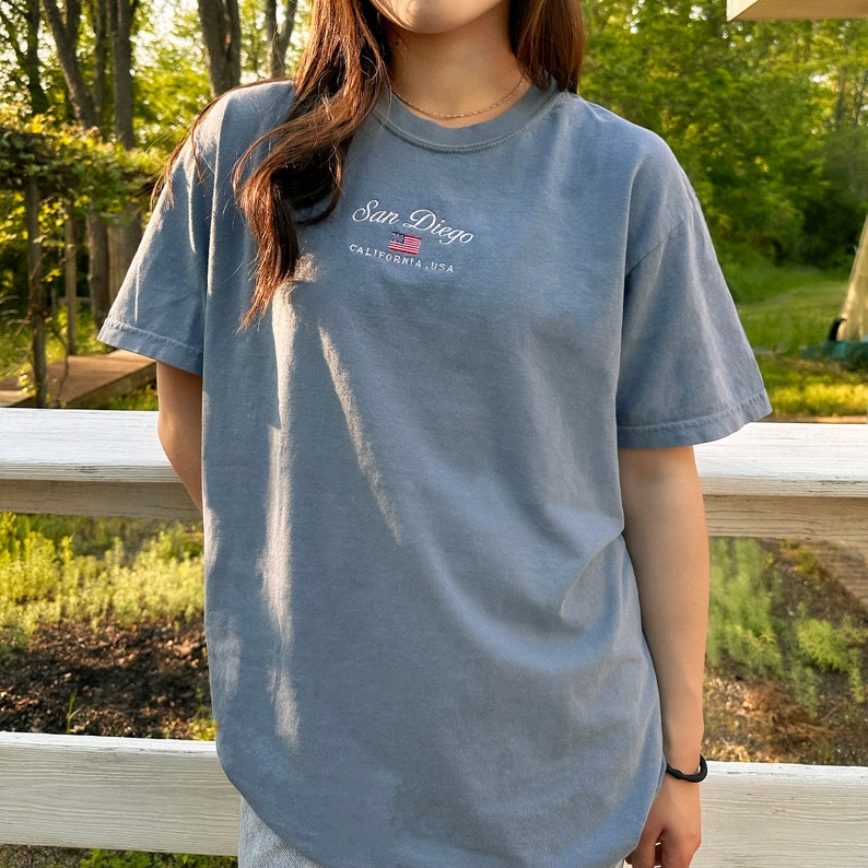 May include: A light blue t-shirt with a white embroidered design that says "San Diego, California, USA" with a small American flag graphic.