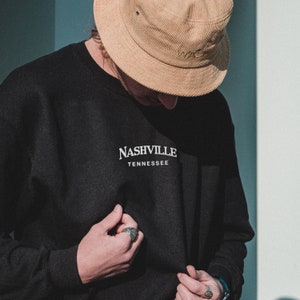 May include: A black sweatshirt with the text "NASHVILLE TENNESSEE" in white lettering. The person is wearing a brown corduroy bucket hat.
