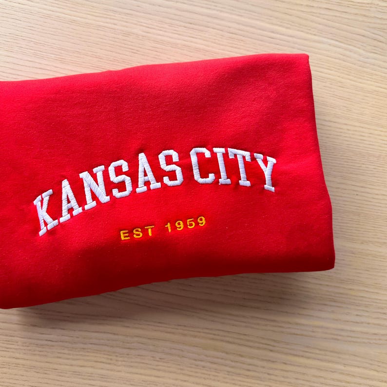 Puede incluir: Una sudadera roja doblada con el texto bordado "KANSAS CITY" en blanco, arqueado sobre el pecho. Debajo del texto est&aacute; "EST 1959" en dorado. La sudadera est&aacute; hecha de un material suave.