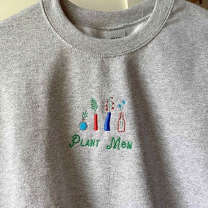 May include: Gray sweatshirt with embroidered green text that reads "Plant Mom" and a design of five plants in vases. The vases are red, blue, and clear.