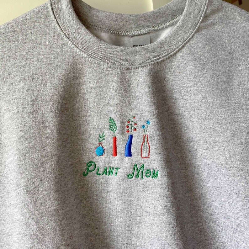 May include: Gray sweatshirt with embroidered green text that reads "Plant Mom" and a design of five plants in vases. The vases are red, blue, and clear.