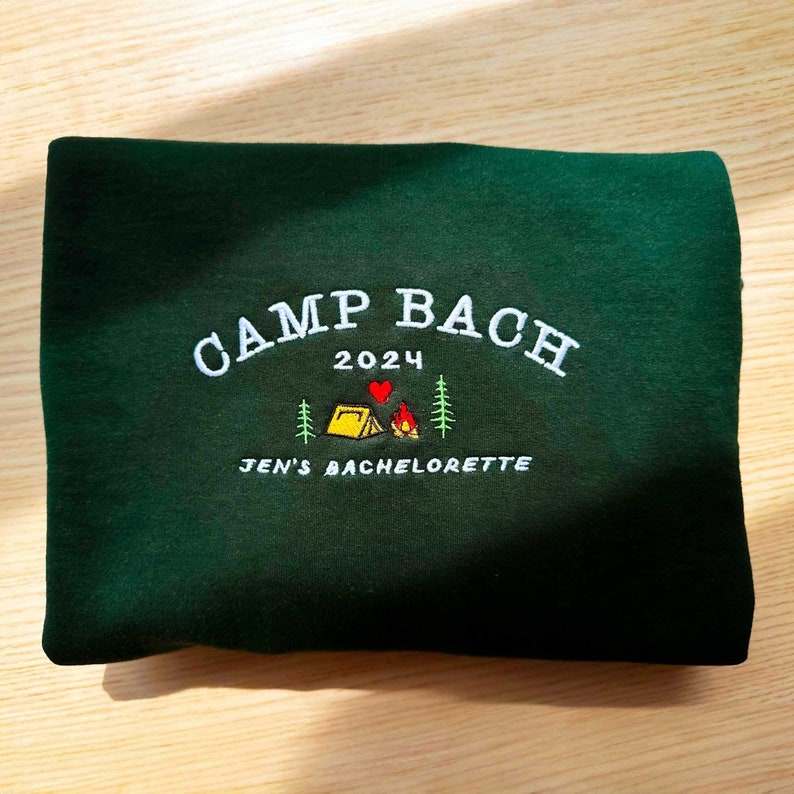 May include: Green sweatshirt with white embroidery that says "Camp Bach 2024" with a red heart and a small embroidered image of a yellow tent, a campfire, and two pine trees. The text "Jen's Bachelorette" is also embroidered below the main text.