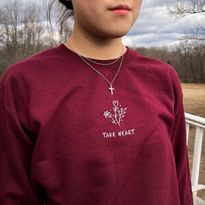 May include: A maroon long-sleeved sweatshirt with a white embroidered design of a flower and the text "TAKE HEART". The model is wearing a silver necklace with a cross pendant.