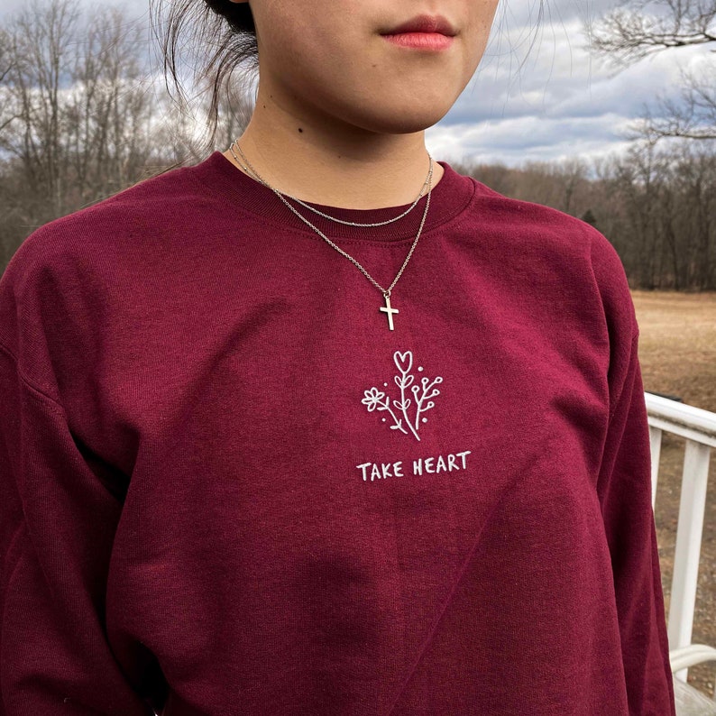 May include: A maroon long-sleeved sweatshirt with a white embroidered design of a flower and the text "TAKE HEART". The model is wearing a silver necklace with a cross pendant.