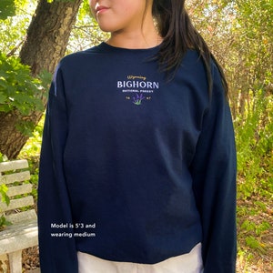 May include: Navy blue long-sleeved sweatshirt with the text "Wyoming Bighorn National Forest" and a small graphic of purple flowers. The model is 5'3" and wearing a medium.