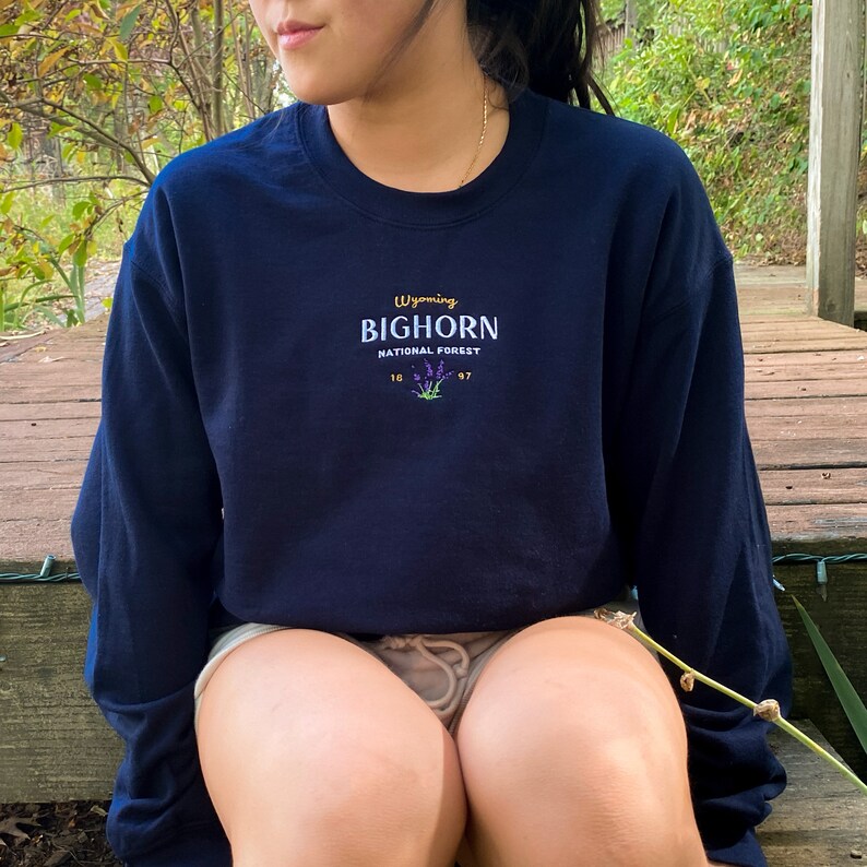 May include: A navy blue long-sleeved sweatshirt with an embroidered design that says "Wyoming Bighorn National Forest" with a purple flower graphic.
