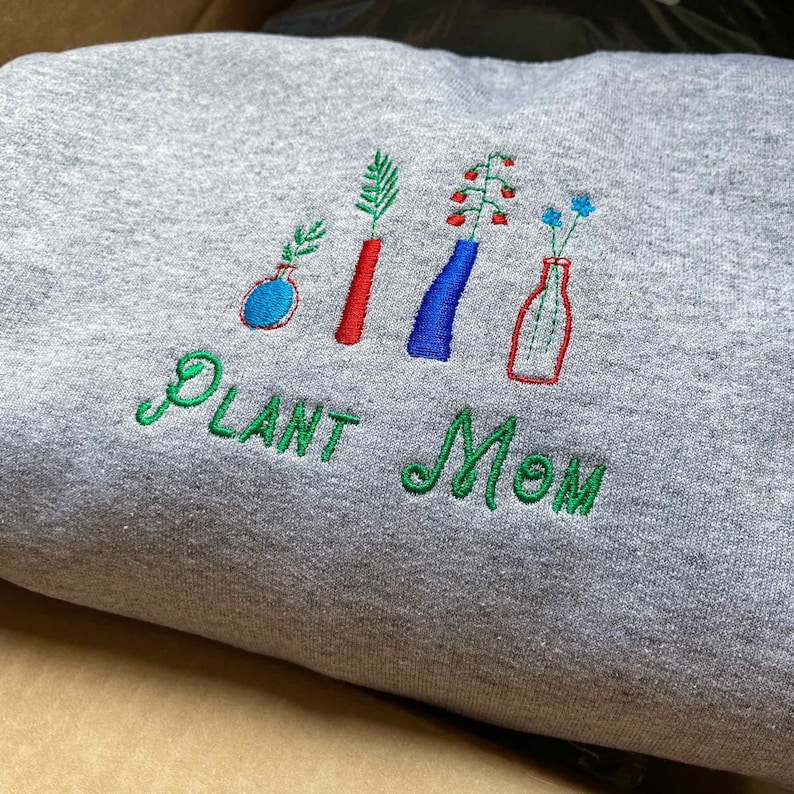 May include: Gray sweatshirt with green embroidered text "PLANT MOM" and five embroidered plants in vases. The vases are red, blue, and clear glass.