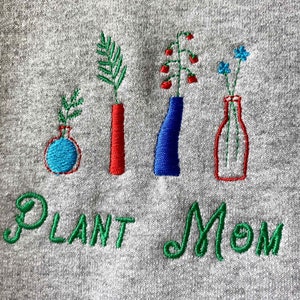 May include: Embroidered gray sweatshirt with the text "Plant Mom" in green thread. The design features four embroidered vases with different colored flowers and plants.