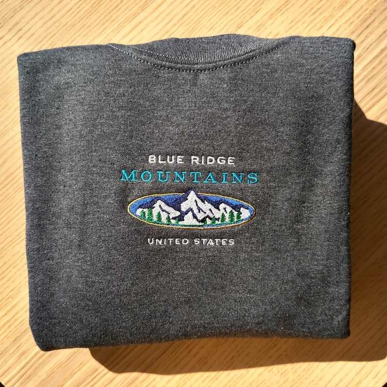 Op de afbeelding: Een donkergrijs sweatshirt met een geborduurd logo met de tekst "Blue Ridge Mountains United States". Het logo toont een blauwe bergketen met groene bomen.