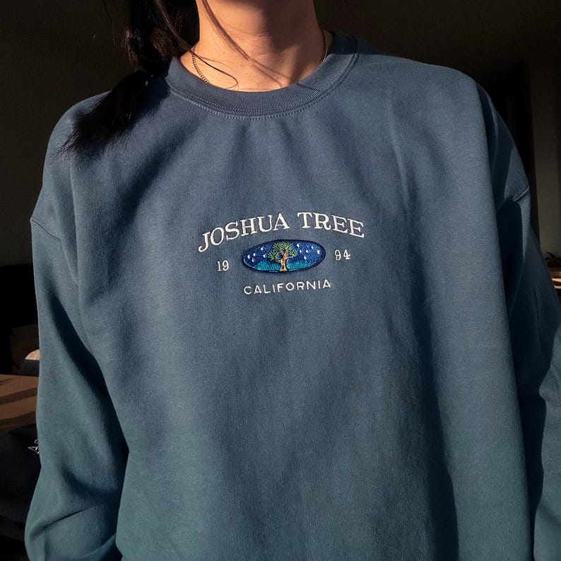 May include: A blue sweatshirt with a white embroidered logo that says "Joshua Tree 1994 California" with a stylized tree and stars inside a circle.