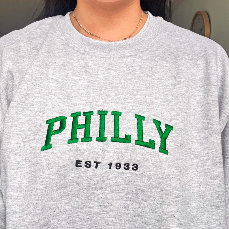 May include: A gray sweatshirt with the word "PHILLY" embroidered in green block letters. Below the word "PHILLY" is the text "EST 1933" in black. The sweatshirt is a crew neck style.