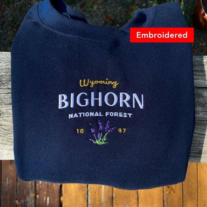 May include: Navy blue sweatshirt with embroidered text that reads "Wyoming BIGHORN NATIONAL FOREST 18 97" with a purple flower graphic.