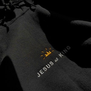 以下が含まれることがあります： 黒いフード付きスウェットシャツに、「JESUS is KING」という白い刺繍のデザインが施されています。KINGの文字の上に金の王冠があります。