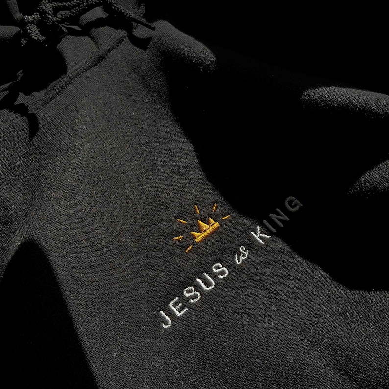 以下が含まれることがあります： 黒いフード付きスウェットシャツに、「JESUS is KING」という白い刺繍のデザインが施されています。KINGの文字の上に金の王冠があります。