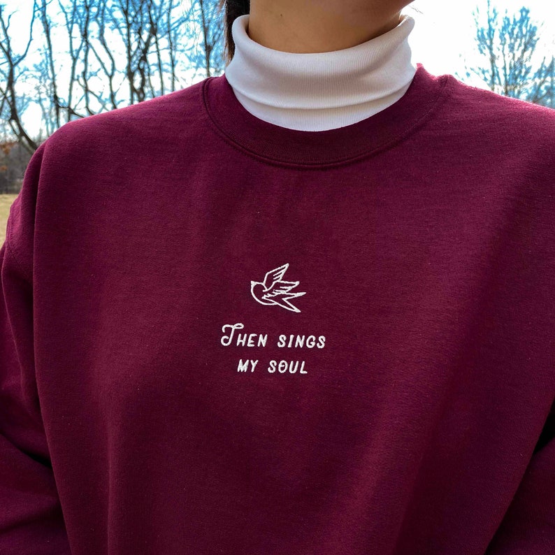 Puede incluir: Una sudadera color burdeos con un dise&ntilde;o bordado en blanco de un p&aacute;jaro volando con el texto "Then Sings My Soul".