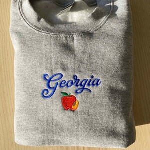 K&ouml;nnte beinhalten: Grauer Pullover mit blau gesticktem Text "Georgia" und einem roten und orangenen Pfirsich-Design.